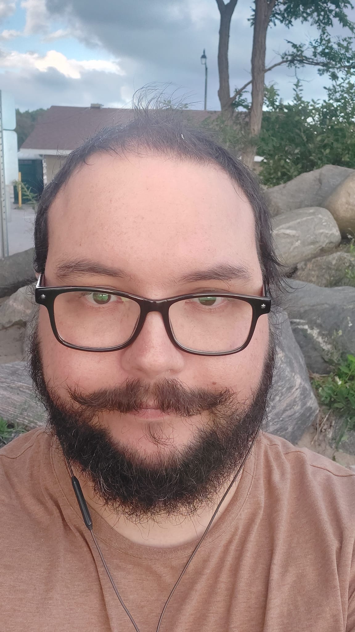 A man short hair and bushy facial hair sits on a rock with more rocks and a small building in the background